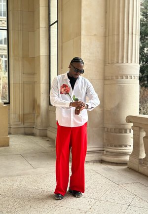 Camisa blanca abotonada con ilustración de rosa roja, combinada con pantalones rojos de pierna ancha y calzado negro. Se llevan gafas de sol y reloj.