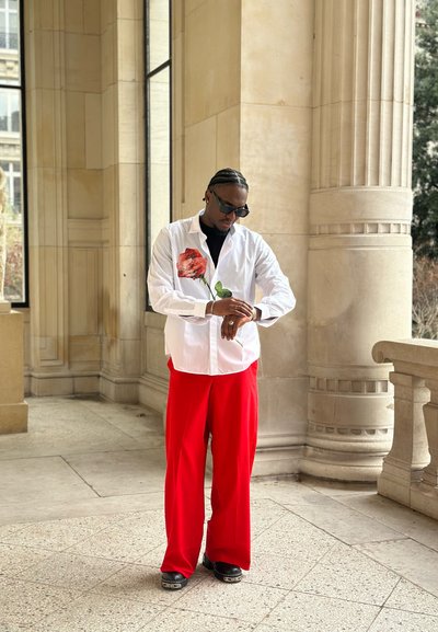 Camisa blanca abotonada con ilustración de rosa roja, combinada con pantalones rojos de pierna ancha y calzado negro. Se llevan gafas de sol y reloj.