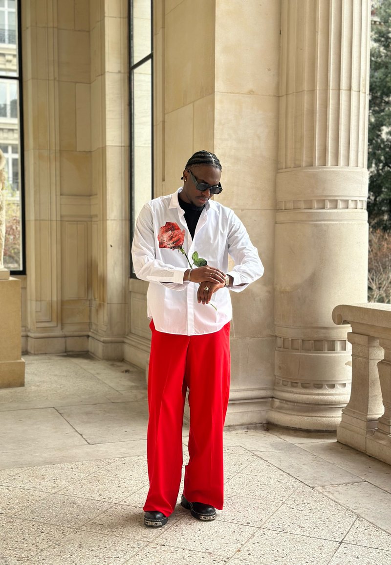 Wit overhemd met knopen en illustratie van een rode roos, gecombineerd met rode wijde broekspijpen en zwart schoeisel. Zonnebril en horloge worden gedragen.