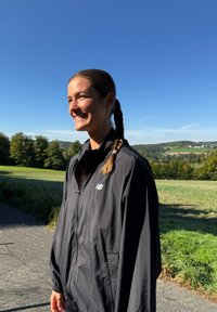 Chaqueta ligera negra con cierre completo y dos bolsillos laterales, que presenta un pequeño logo blanco en el lado izquierdo del pecho, usada al aire libre contra un cielo despejado.