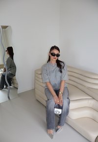 Grijze oversized t-shirt met gedrukte tekst, gecombineerd met grijze jeans, metallic zilveren pointed schoenen en een kleine zilveren handtas, op een gebogen beige bank.