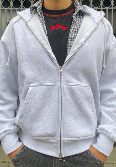 Sudadera de cremallera de color gris claro en tejido suave, con dos bolsillos frontales, cordones y una camiseta negra de contraste por debajo con estrellas rojas.
