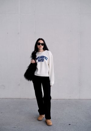 Mujer con gafas de sol negras, sudadera blanca con texto azul, pantalones negros, botas marrón claro, llevando un bolso negro de pelo, frente a una pared gris lisa.