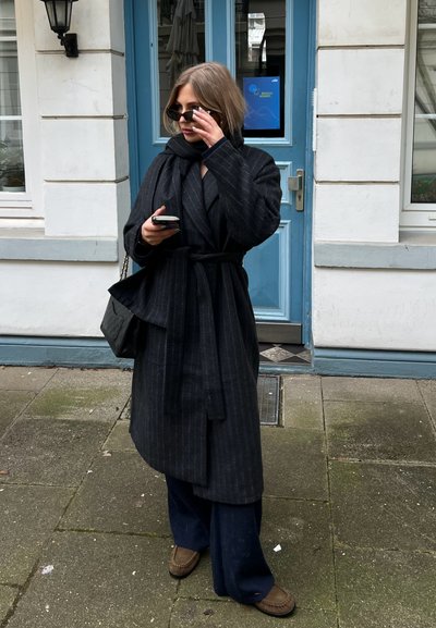 Abrigo de rayas negras con cinturón atado, usado sobre un conjunto oscuro. Bolso negro, gafas de sol, sosteniendo un teléfono. Fondo de puerta azul clara.
