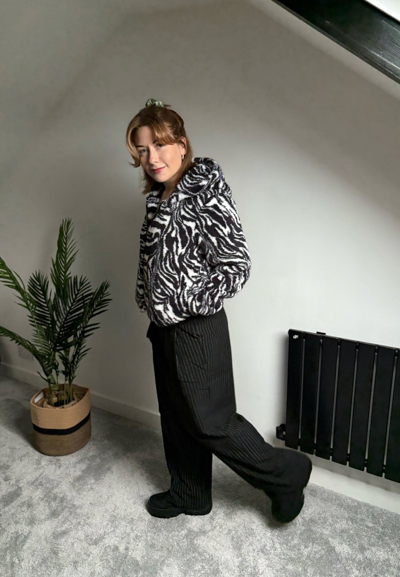 Jeune personne portant une veste à motif zèbre et un pantalon noir, posant à l'intérieur près d'une plante en pot et d'un radiateur noir sur un tapis gris clair.