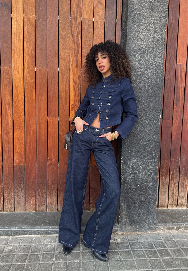 Femme aux cheveux bouclés appuyée contre un mur en bois et béton, portant une veste de style militaire bleu foncé, un jean à jambes larges et des chaussures noires.