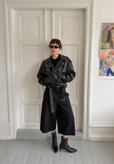 Persona con chaqueta de cuero negra, pantalones negros de pierna ancha, botas negras con tacón y gafas de sol oscuras, de pie en el interior frente a puertas blancas con paneles.