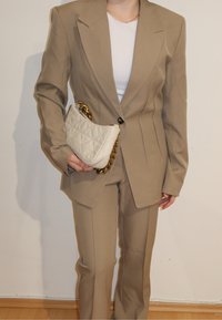 Personne portant un costume beige ajusté et un haut blanc, tenant un petit sac matelassé blanc avec une anse chaîne dorée.