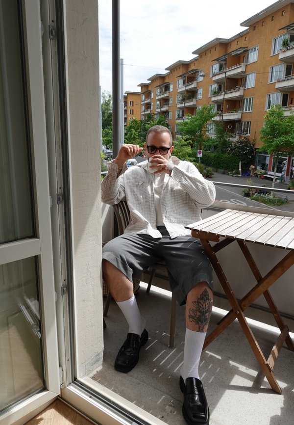 Muž s slunečními brýlemi a světlou košilí sedí na balkóně a pije z bílého hrnku, s viditelným tetováním na noze a panelovými domy v pozadí.