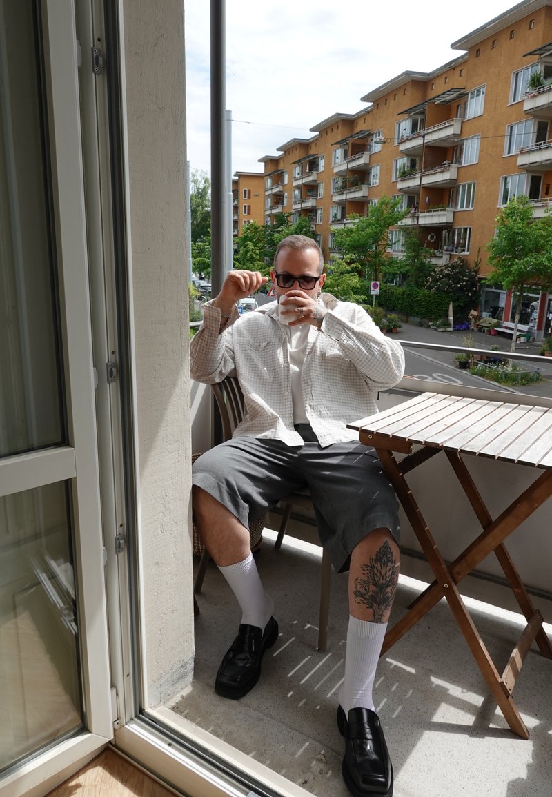 Mann mit Sonnenbrille und hellem Hemd sitzt auf dem Balkon und trinkt aus einer weißen Tasse, mit sichtbarem Tattoo am Bein und Wohnhäusern im Hintergrund.
