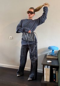 Femme portant un sweat-shirt sombre surdimensionné, un pantalon de jogging noir, de grandes lunettes de soleil, tenant un sac noir et remontant sa queue de cheval à l'intérieur près d'une étagère.