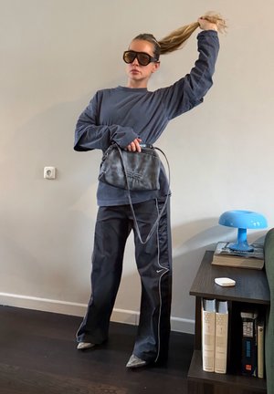 Mujer con sudadera oscura de gran tamaño, pantalones deportivos negros, gafas de sol grandes, sosteniendo un bolso negro y levantando su coleta en el interior junto a una estantería.