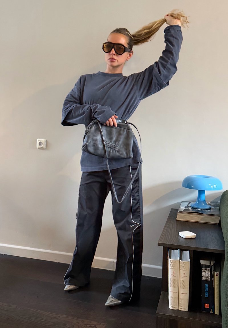 Femme portant un sweat-shirt sombre surdimensionné, un pantalon de survêtement noir, de grandes lunettes de soleil, tenant un sac noir et relevant sa queue de cheval à l'intérieur près d'une étagère.