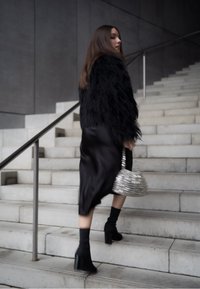 Casaco de pelúcia sintética preto, vestido midi de cetim preto, bolsa metálica prateada, meias de tornozelo pretas e botas de salto. Escadas ao fundo.