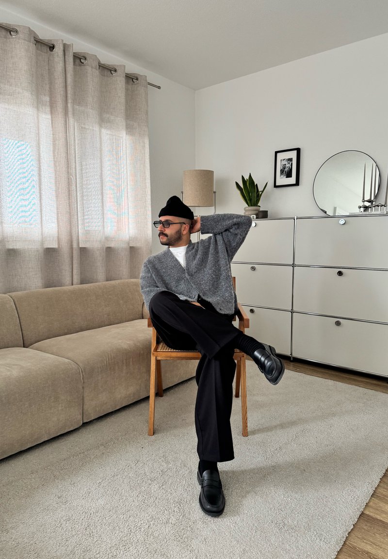 Man in glasses, black beanie, gray sweater, black pants, and shoes sitting cross-legged on wooden chair in a modern living room.