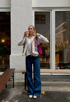 Jersey de punto con bordado floral en el frente, ribete de pelaje sintético color crema; blusa blanca de manga larga; jeans de mezclilla azules de pierna ancha; bolso rosa; sosteniendo una taza de café.