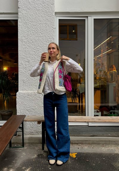 Jersey de punto con bordado floral en el frente, ribete de pelaje sintético color crema; blusa blanca de manga larga; jeans de mezclilla azules de pierna ancha; bolso rosa; sosteniendo una taza de café.