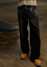 Personne portant un jean foncé et des bottes couleur sable debout sur un bitume mouillé avec des taches de lumière réfléchissante la nuit.