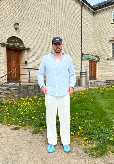 Camisa de botón azul claro, pantalones de lino blancos y zapatillas azules. El fondo presenta un edificio de ladrillo y dientes de león en la hierba.