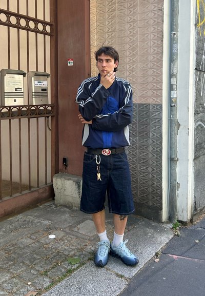 Chaqueta deportiva azul con detalles grises, pantalones cortos de mezclilla y zapatillas deportivas azul claro. Los accesorios incluyen un llavero y un cinturón con una hebilla circular.