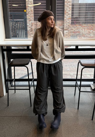 Joven mujer con boina, chaqueta beige, camisa blanca, pantalones grises holgados y botas negras, de pie entre dos taburetes en un interior.