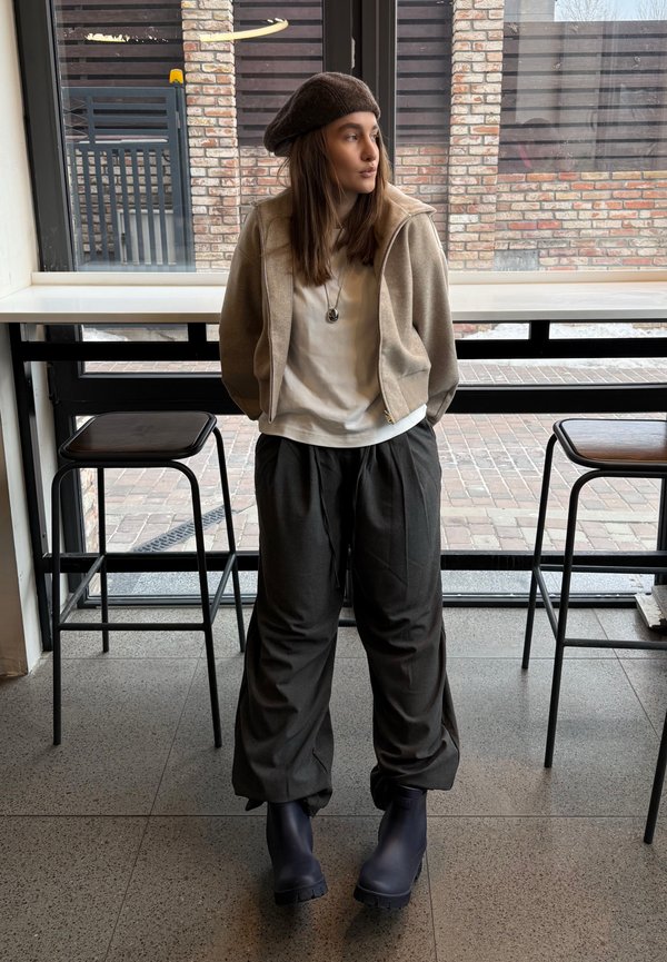 Junge Frau mit Baskenmütze, beiger Jacke, weißem Hemd, lockeren grauen Hosen und schwarzen Stiefeln, die drinnen zwischen zwei Barhockern steht.