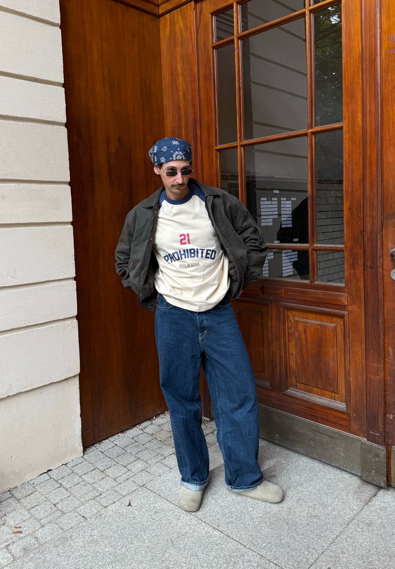Legeres Outfit mit einem cremefarbenen T-Shirt mit rotem und blauem Schriftzug, grüner Bomberjacke, dunkelblauen Jeans mit weitem Bein und grauen Clogs.