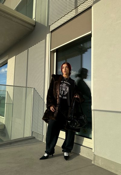 Abrigo de piel sintética marrón sobre una camiseta gráfica negra, pantalones negros holgados y tacones negros de punta con detalles blancos, de pie junto a una ventana.