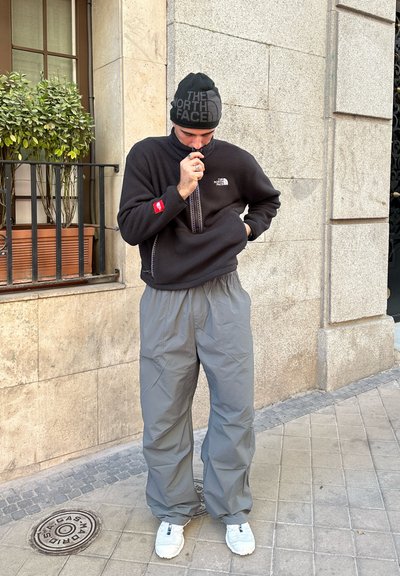 Sudadera de forro polar negra con cremallera de cuarto, pantalones impermeables grises y zapatillas blancas. Incluye un gorro negro con un patrón de logotipos.
