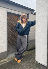 Jeune personne appuyée contre un mur blanc, portant un chapeau doublé de fourrure, une veste bleue, un pantalon large gris et des bottes beige sur un chemin pavé.