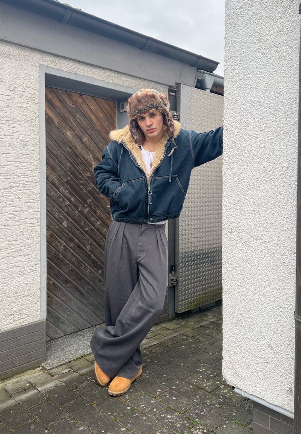 Jeune personne appuyée contre un mur blanc, portant un chapeau doublé de fourrure, une veste bleue, un pantalon gris large et des bottes beige sur un chemin pavé.