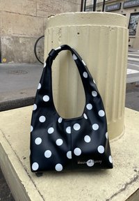 Sac à main en cuir noir à pois blancs avec le logo "House of Sunny", posé sur un élément urbain beige près d'un passage piéton.