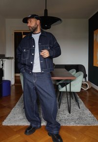 Jeansjack met knopenfront, twee borstzakken en contrasterende stiksels, gecombineerd met losse denim broek en zwarte schoeisel.