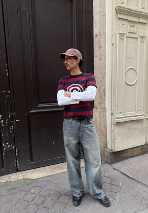 Hombre con camiseta de rayas, gorra, gafas y vaqueros holgados, de pie con los brazos cruzados en una calle de la ciudad cerca de una puerta oscura y una fachada de edificio clara.