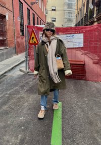 Capa longa verde oliva, cachecol cinza claro, jeans azuis, sapatos bege. Pequena bolsa bege. O fundo mostra barreiras de construção e sinais de trânsito.