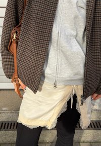 Personne portant un legging noir, une jupe en dentelle beige par-dessus, un sweat à capuche gris zippé, une veste marron à carreaux et tenant un sac à main en cuir marron.