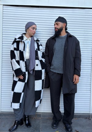 Abrigo de tablero de ajedrez en blanco y negro, top gris, bufanda gris y pantalones anchos gris. Pantalones negros con rayas finas y botas negras lisas.