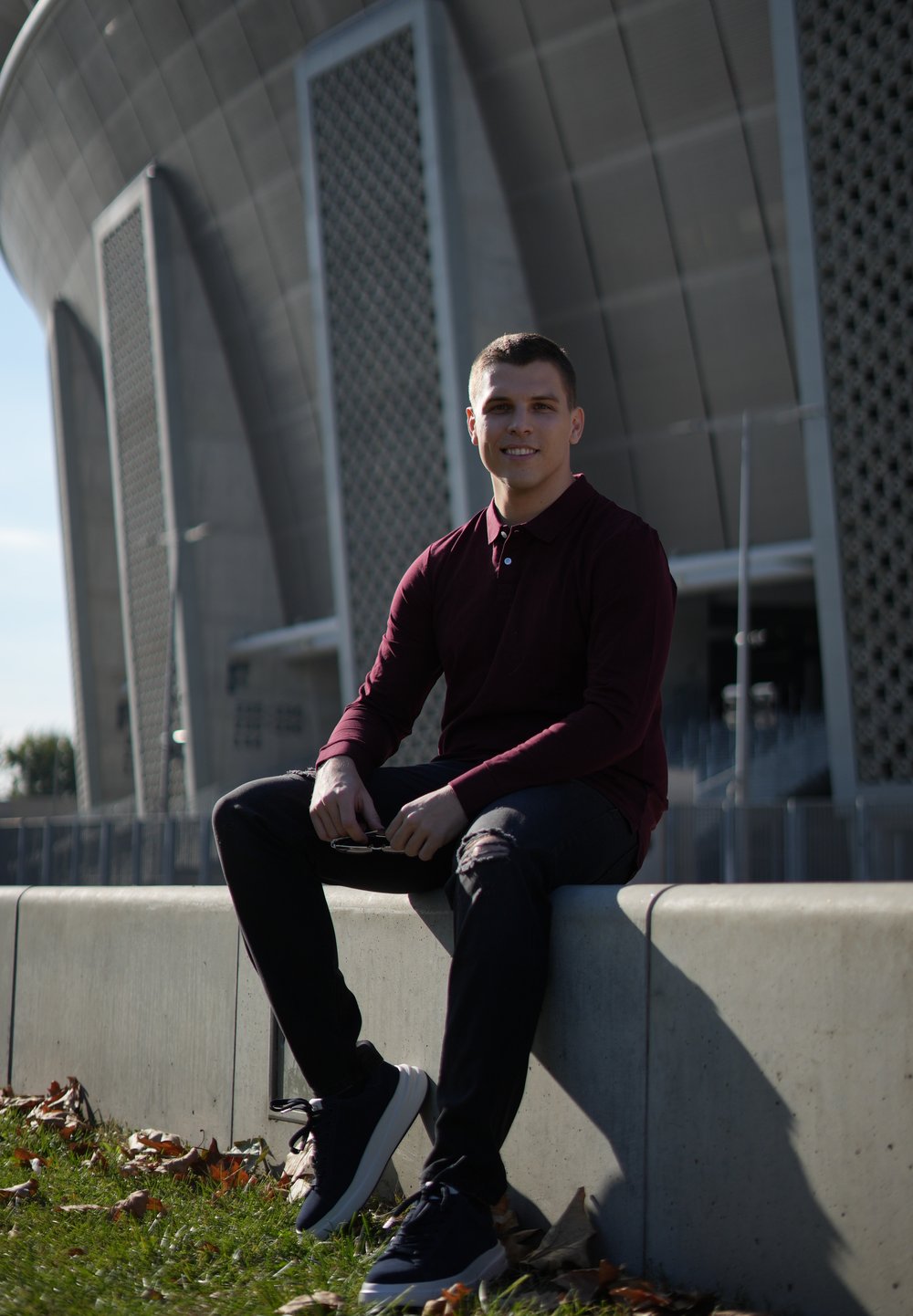 Young man in a burgundy long-sleeve shirt and black jeans sitting on a grey concrete wall, wearing dark sneakers. Industrial building in background.