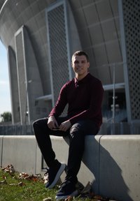 Young man in a burgundy long-sleeve shirt and black jeans sitting on a grey concrete wall, wearing dark sneakers. Industrial building in background.