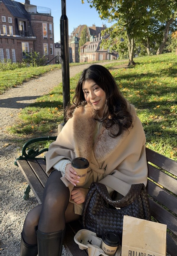 Cappotto beige con colletto di pelliccia, leggings scuri, stivali neri alti, tiene una tazza di caffè, seduta su una panchina in un parco con vegetazione sullo sfondo.