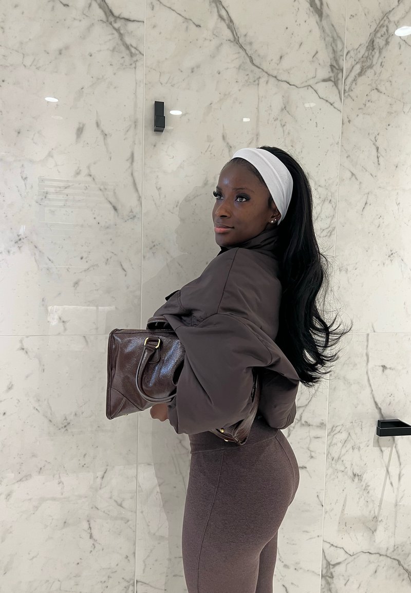 Femme aux cheveux longs avec un bandeau blanc, portant une tenue marron, tenant un sac à main en cuir foncé, debout contre un mur en marbre à l'intérieur.