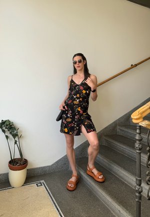 Vestido negro con estampado floral y tirantes finos, con una abertura lateral. Sandalias de plataforma marrones. Los accesorios incluyen gafas de sol y un bolso cruzado negro.