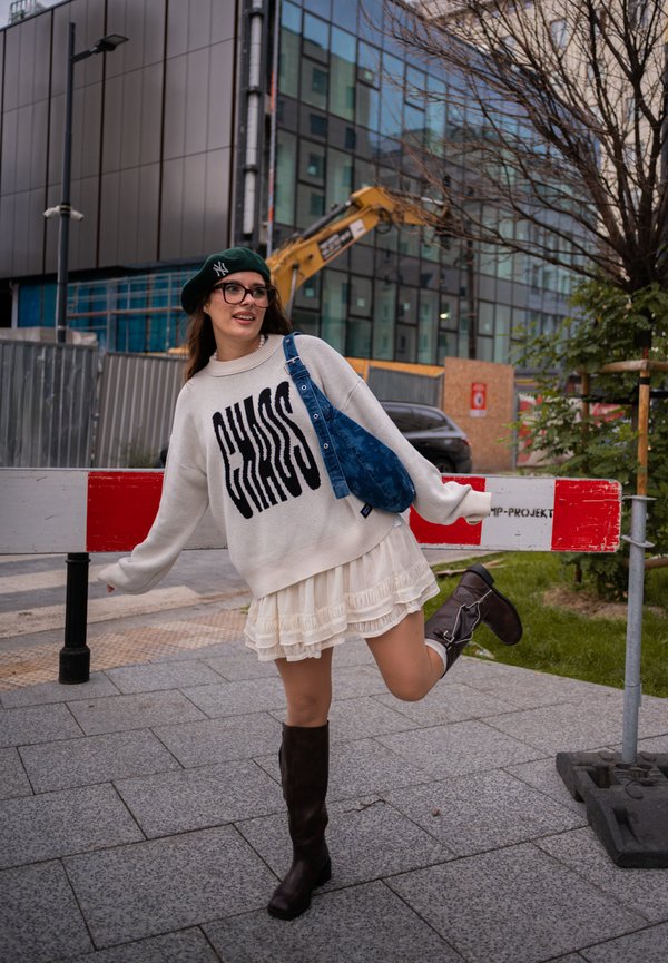Joven mujer con un suéter blanco con texto "CHAOS", falda blanca, botas negras y boina verde posando juguetonamente en la acera urbana cerca de una obra en construcción.