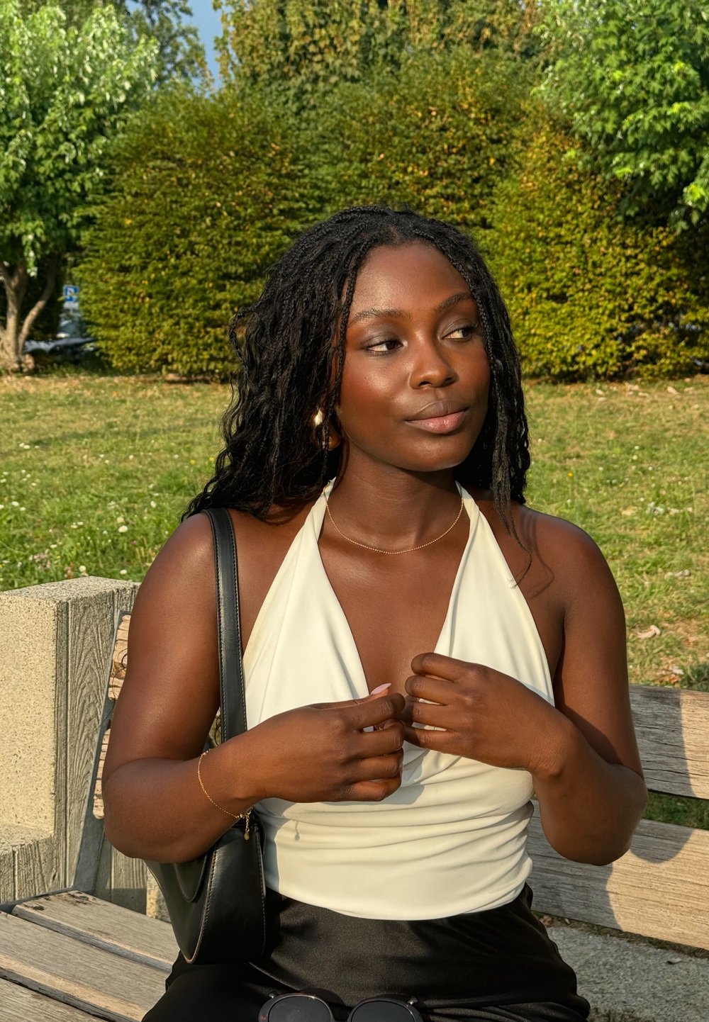 Une femme portant un haut blanc sans manches ajuste son haut en étant assise sur un banc en bois. Elle a des cheveux noirs texturés et un sac à main noir. Un feuillage vert l'entoure.