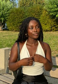 Une femme portant un haut blanc sans manches ajuste son haut en étant assise sur un banc en bois. Elle a des cheveux noirs texturés et un sac à main noir. Un feuillage vert l'entoure.