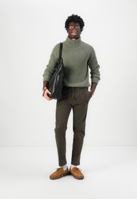 Jersey de canalé verde oliva de cuello alto, pantalones marrones de corte ajustado, zapatos de cordones en tono marrón claro y un bolso de hombro de piel negra. Fondo neutro.