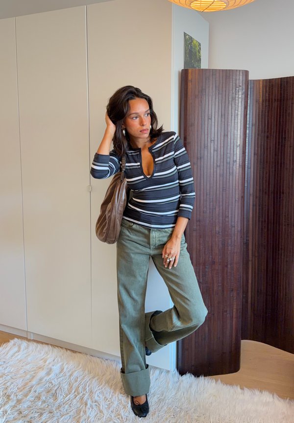 Mujer de pie en el interior vestida con una camiseta de manga larga a rayas, pantalones sueltos con puños, zapatos negros y un bolso marrón al hombro, apoyada contra una pared blanca.