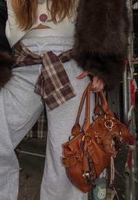 Smeđa kožna torba s više metalnih detalja, privjesaka i remenom. Mekani, teksturirani završetak; drži je osoba u sivim trenirkama.