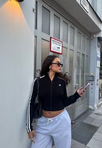 Fekete bordás, rövid fazonú, cipzáras pulóver fehér csíkokkal, szürke melegítőnadrág és napszemüveg viselésével. Telefont tart a kezében, és egy épület mellett áll, amelynek felirata van.