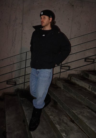 Joven con chaqueta negra, jeans azules y gorra negra apoyado en una barandilla junto a unas escaleras de concreto por la noche.